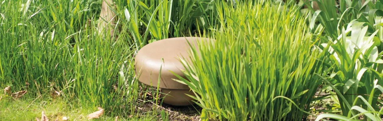 Monitor Audio CLG-W12 – subwoofer completamente interrato nel giardino, solo la porta a cupola visibile tra il prato, finitura marrone